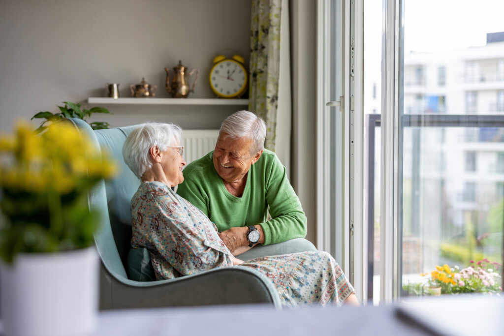 An elderly couple sitting in a chair, gazing out the window, holding hands, with a serene expression on their faces.