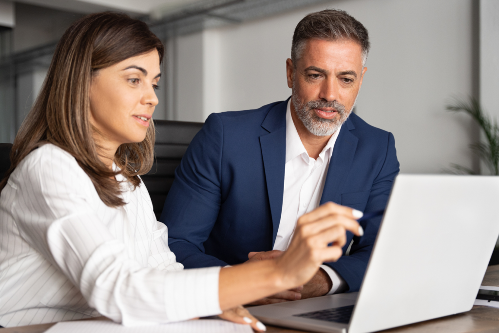 Team of diverse partners - mature business man and business woman discussing project on laptop sitting at table in corporate office.