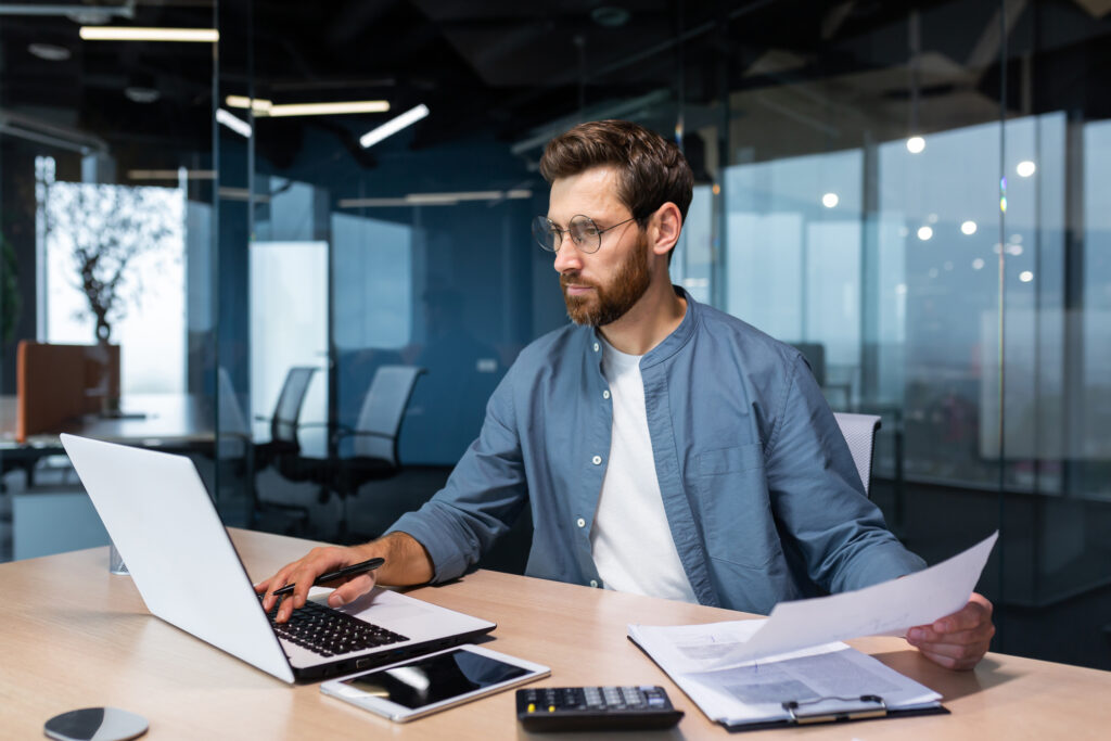 A serious young man accountant, financier, analyst, auditor sits in the office at the table. He holds documents and a pen in his hands, checks accounts, finances, types on a laptop. business office worker