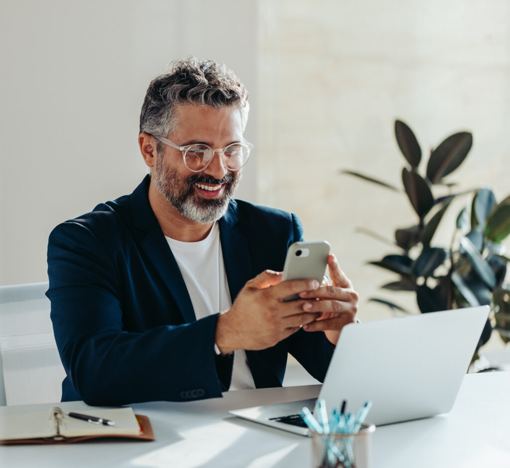 Cheerful, mature professional man is engrossed in his smartphone while sitting at a white desk in a sunny, contemporary office space. His laptop and notepad are at hand, suggesting productivity and connectivity.